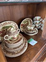 Shelf showing stacked dinnerware plates, cups, and saucers with rural scene patterns in brown and green.