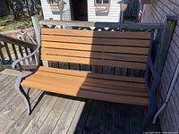 Full view of the bench on a wooden deck. Shows wrought iron frame and wooden slat seating and backrest.