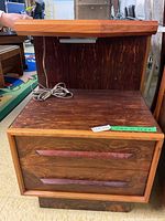 Front and side view of one wooden bedside table showing two drawers with carved wooden handles and a built-in shelf above with an attached electrical cord, highlighting wood grain and finish.