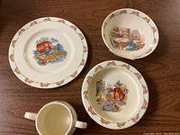 Four-piece Bunnykins children's dish set on wooden table including a plate, two bowls, and one handled cup. All pieces have bunny character illustrations and bunny motif borders.
