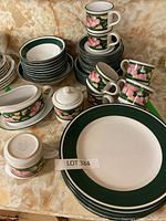 Full set of vintage porcelain dinnerware with green border and pink floral pattern, including plates, cups, and serving pieces, placed on a beige marble countertop.