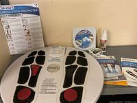 Overview of Dr. Ho’s Circulation Promoter device showing the foot base with black textured pads and red circular areas, connected wire, Quick Start Guide, manuals, DVD, and Pain-Aid spray bottle.