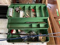 Photo of open green metal fishing tackle box showing multiple compartments with lures and some fishing gear inside, and fishing rods placed across the box.