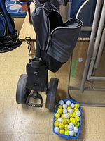 Photo showing black golf push cart with wheels and handle next to basket of white and yellow golf balls.