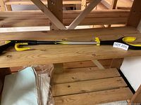 Full length photo of the Handi-Grip Pro grab assist tool showing yellow and black handle, long metal shaft, and grabber claw end, placed on a wooden surface.