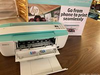 Front view of HP DeskJet 3755 printer with front compartment open exposing ink cartridge slots. The packaging box is visible in background advertising phone-to-print capability.