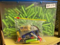 Plastic container filled with numerous green foam darts with blue suction cup tips. In front, a sealed clear plastic bag contains Nerf accessories and some foam darts, labeled with '250 CRAZY BALLS'.