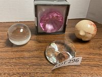Four paperweights on a wooden table: clear dome crystal, faceted clear crystal, faceted pink crystal in box, and round marble paperweight.