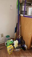 Corner with multiple brooms and cleaning tools, some supplies on the floor and shelf