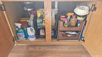 Cabinet under sink with multiple cleaning products arranged inside, including sprays, bleach, and pads.