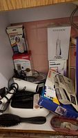 Photo showing various healthcare and personal care items including boxed oral irrigators, clothes shaver, blood pressure monitor, hairdryer, hairbrush, scale, and clipper attachments.