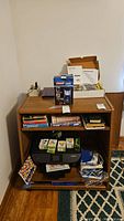 Wooden shelving unit filled with various items including books, printer, boxed router, ink cartridges, and cables.