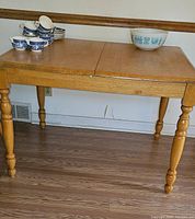Light wood kitchen table with turned legs. Leaf stored under table, visible as the table has a center split for leaf insertion.
