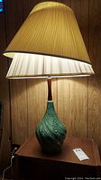 Mid-century pottery lamp with green textured base and olive green pleated shade shown lit on a wooden surface.