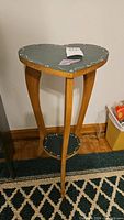 Side view of the wooden plant stand showing heart shaped top painted dark green with decorative gold edging, natural wood legs with curved design.