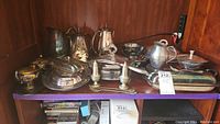 Complete set of various silver plated serving and tea pieces displayed on a wooden shelf. Includes tea pots, serving trays, bowls, and weighted sterling salt and pepper shakers.