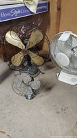 Photo of two fans on concrete floor including a vintage Mastercraft metal desk fan and a modern white Lasko table fan.