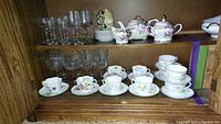 Shelf displaying hand painted teapot, creamer, sugar bowl, surrounding hand painted teacups and saucers, and etched glass stemware above and beside.
