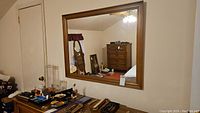 Wood framed wall mirror hung on wall, reflecting part of bedroom and dresser. Shows brown wood frame with simple molding details.