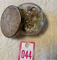 Clear glass trinket dish with floral cushioned lid filled with various jewelry including pearl beads, pins, and purple stone pieces.