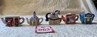 Five miniature teapot figurines made by Wade for Red Rose Tea, arranged in a row on a light beige fabric surface. Figurines include a fire truck, church, rocking horse, a detailed teapot, and a blue teapot with writing.
