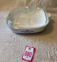 Top angled view of vintage Corning Ware 10-inch blue cornflower casserole dish with glass lid on beige fabric background.