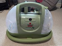 Front view of Bissell Little Green ProHeat upholstery cleaner showing power button, clear water tanks, and coiled hose at base.