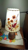 Front view of vintage lamp showing parchment shade with pressed floral and leaf design and wooden base on a cloth with colorful print.
