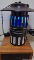 Front view of black cylindrical Dynatrap insect trap showing blue UV light inside and mesh lower section with Dynatrap branding.