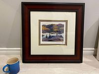 Framed Franklin Carmichael limited edition print showing a landscape with hills and trees in various colors, next to a cup for size reference.
