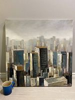 Front view of the acrylic painting showing a stylized city skyline with tall buildings and muted color palette. A coffee mug is included for scale.