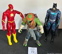 Three action figures standing: The Flash, Michelangelo TMNT, and Batman, showing full view and size scale