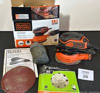 Photo showing the Black & Decker 5-inch random orbit sander with a red sanding disc attached, dust collection bag connected, extra sanding discs laid out, original box, and instruction manual.