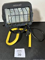 Front view of the Noma work light showing glass lens, protective metal grill, yellow tubular stand, power cord, and plug.