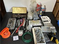 Full assortment of tools and hardware on black surface showing suction cup tool, various fasteners, a wire brush, wrenches, and spray cans