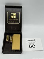 Photo of open black case with gold 'Win International' branding, showing gold-tone lighter and two batteries inside.