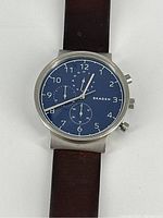 Front view of the watch showing blue dial, chronograph subdials, and stainless steel case with leather strap.