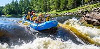 Group of rafters in yellow helmets and life jackets paddling a blue Wilderness Tours raft through whitewater rapids