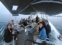 Group of about a dozen guests seated in the cockpit of a sailboat at dusk with wine glasses and cheese plates on the table