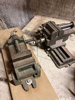 Two metalworking vices placed on a rough wooden plank on a workbench, showing overall condition and design.