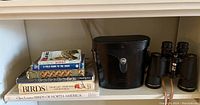 Binoculars with case and six bird books stacked on shelf showing titles on spines