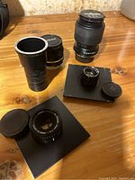 Five camera lenses on wooden surface, showing variety in size and brand caps (Nikon, Tamron). Two lenses mounted on black plates.