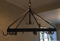 Full view of black wrought iron pot rack hanging from chain in a room with patterned wallpaper and windows.