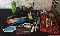 Full view showing assorted kitchen utensils and tools arranged on tabletop, including mandolin, potato masher, garlic press, cheese cutters, nut crackers, and other small items in red containers.