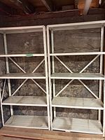 Front view of two white metal shelving units with four shelves each against a stone wall. Shelves appear used and dirty.
