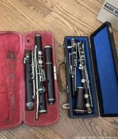 Two vintage clarinets disassembled into sections placed in separate open cases on a wooden floor; one case lined with blue fabric, the other with red velvet.