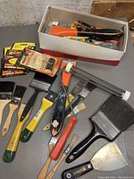 Photo showing assortment of paintbrushes, scrapers, staples, and paint roller in and around a cardboard box.