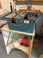 Front and side view of the green pneumatic assembler mounted on shelf, showing dual aluminum work surfaces and red air hoses