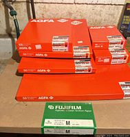 Multiple boxes of Agfa Multicontrast Premium MCP 310 black and white resin-coated photo papers in bright orange packaging with sticker labels showing sheet counts and sizes, and one green box of Fujifilm Crystal Archive color photo paper.