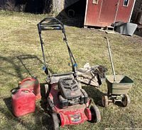 Full view of the lawnmower with grass catcher bag, two red Jerry cans, and green fertilizer spreader on grassy ground